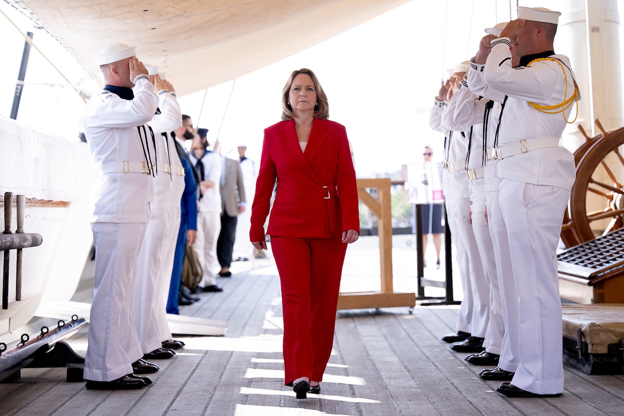 A civilian leader in a red business suit walks a boardwalk flanked by saluting sailors.