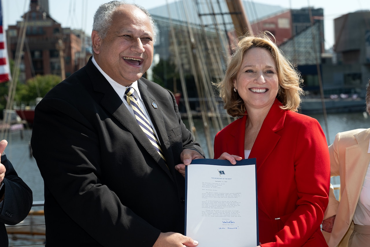 A civilian in a red suit stands next to another in a dark suit, both holding a citation.