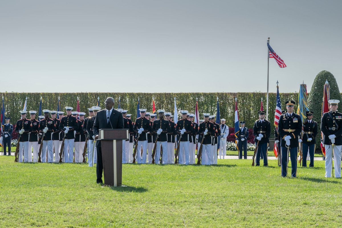 A government official speaks from behind a lectern on a grassy field flanked by service members in uniform and flags.