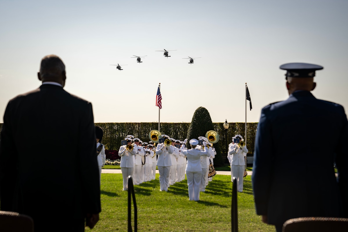Two men face a band performing near two flags as four helicopter fly above.