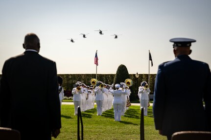 Two men face a band performing near two flags as four helicopter fly above.