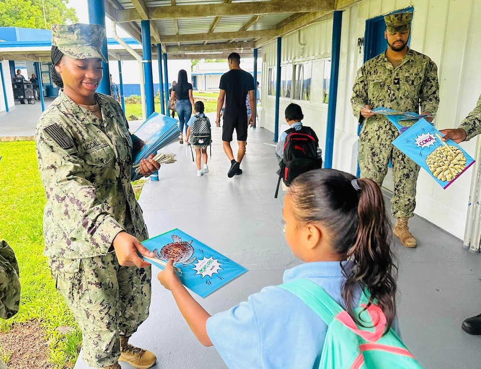 HAGAT, Guam (Aug. 15, 2024) - Seaman Yeoman Charanna McCall hands a folder to a student at the Marcial A. Sablan Elementary campus in Hågat on the first day of school, Aug. 15. McCall and other NBG Administrative Department staff handed out folders and pens. The folders were produced under Commander, Navy Installations Command in support of the NBG Listed Species and Marine Resources Outreach.