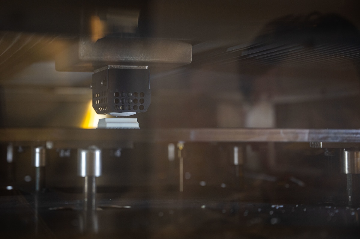 A close-up image of a 3D printer in operation. The image shows the interior of the printer with the printing nozzle and object being printed in focus.