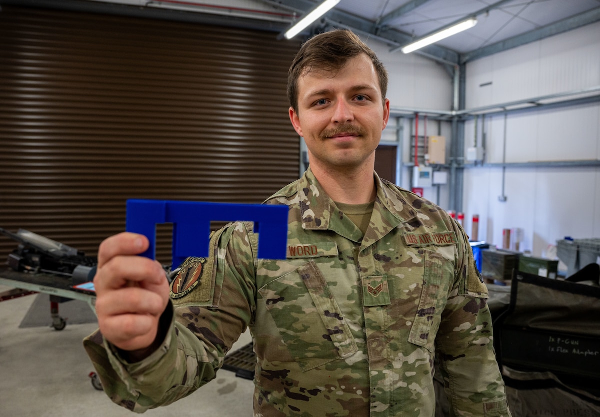 An Airman holds his innovation, a 20 mm ammunition guide insert, in front of a replenisher table connected to an ammunition loader.