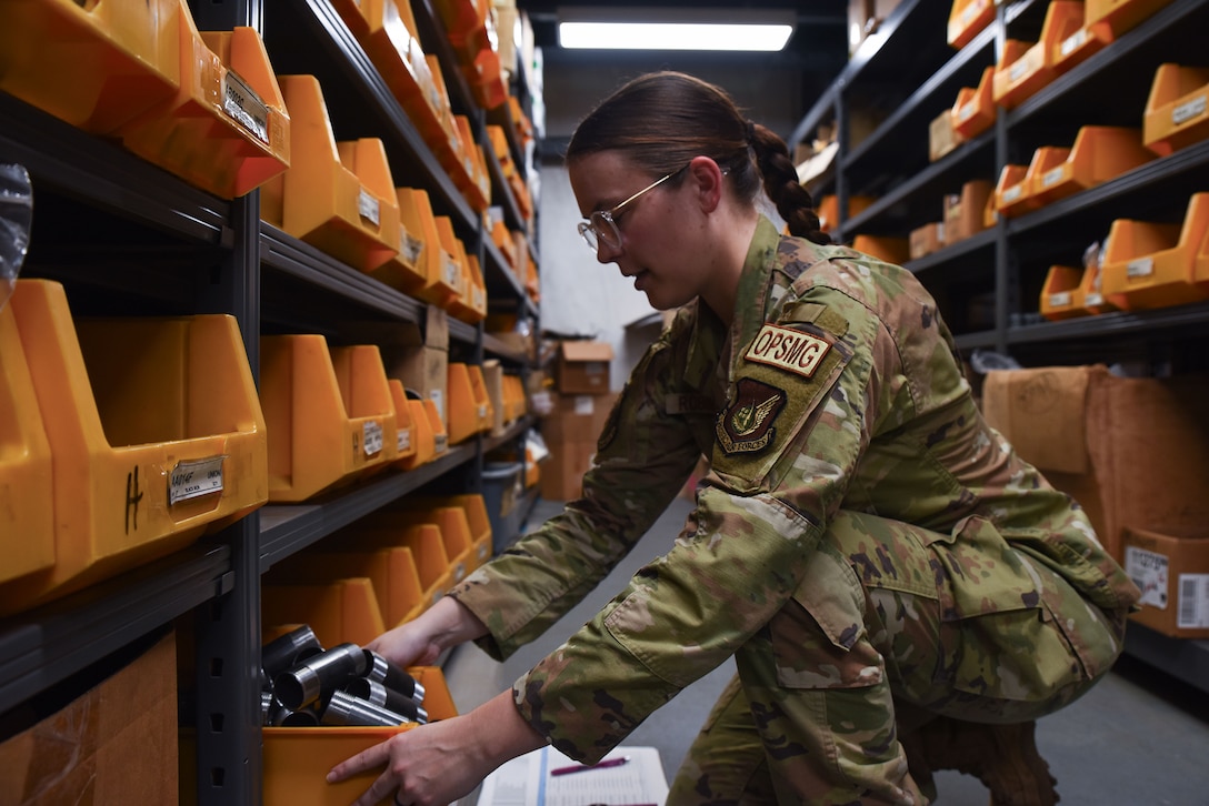 An airman kneels on the floor to count tools in an orange container inside a storage room.