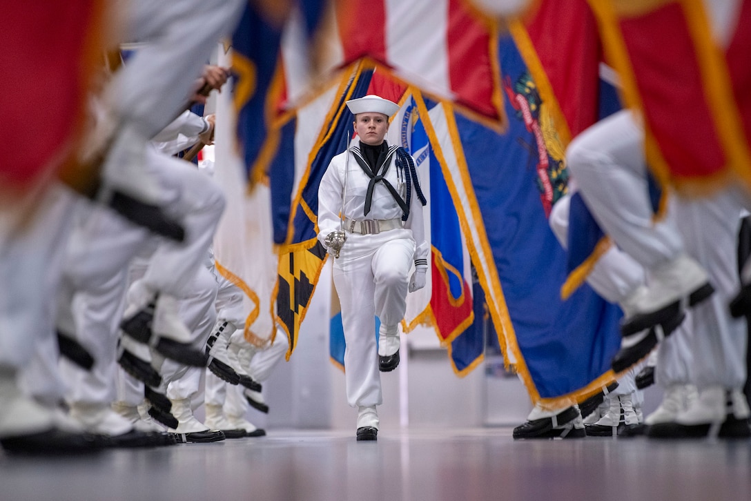 A Navy recruit marches with a ceremonial sword as the feet of flag bearers marching to the left and right are visible in the photo.