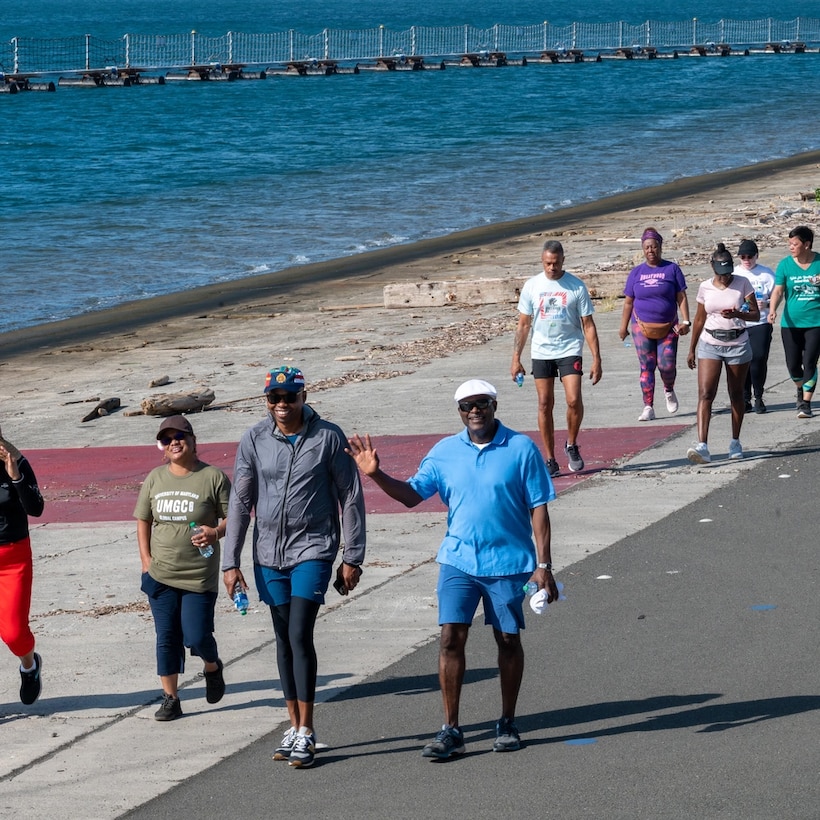 People walking alongside water to the left. Man in blue waving.