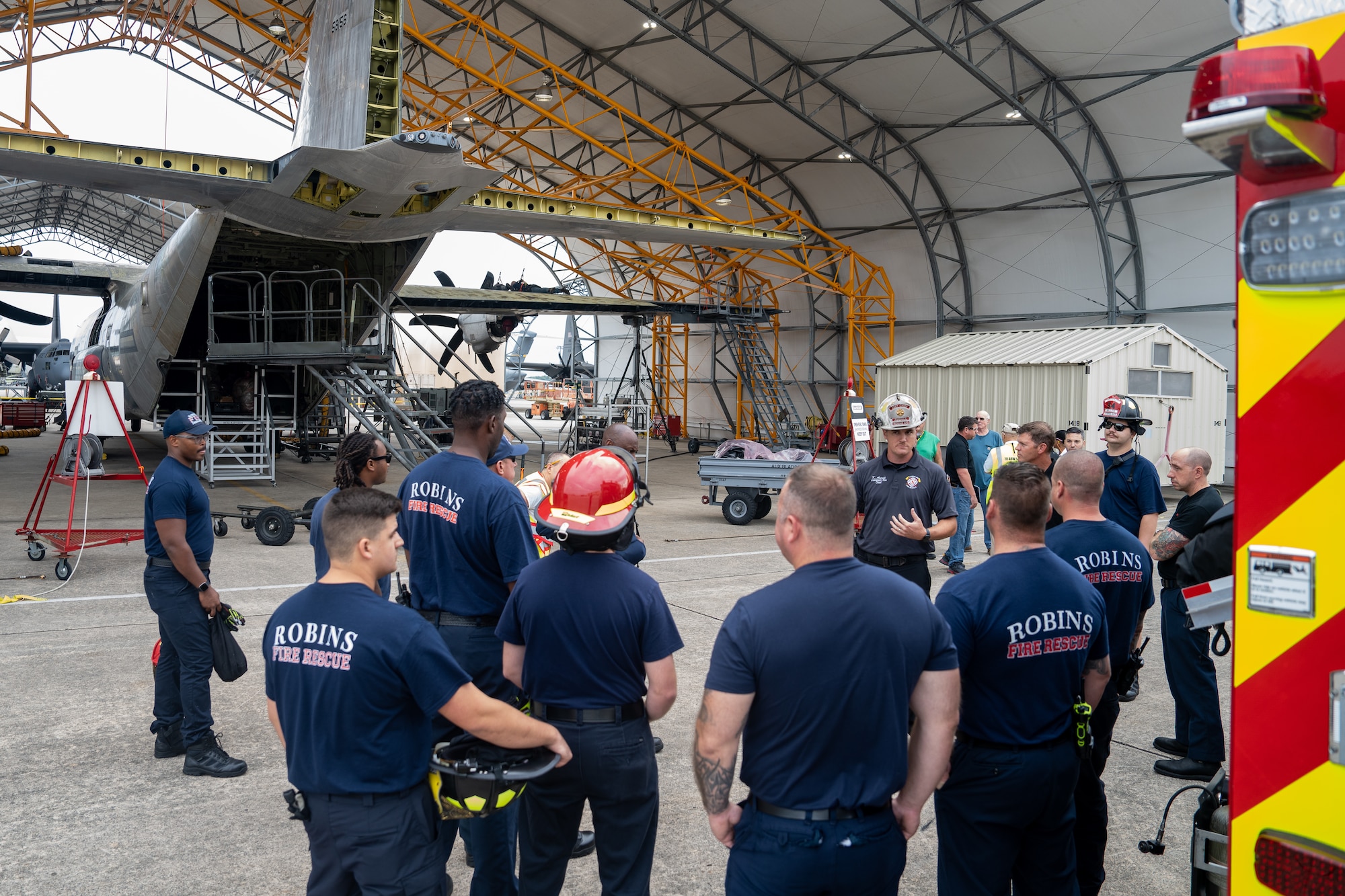 A group of firefighters stand between a fire truck and C-130 Hercules aircraft discussing upcoming training