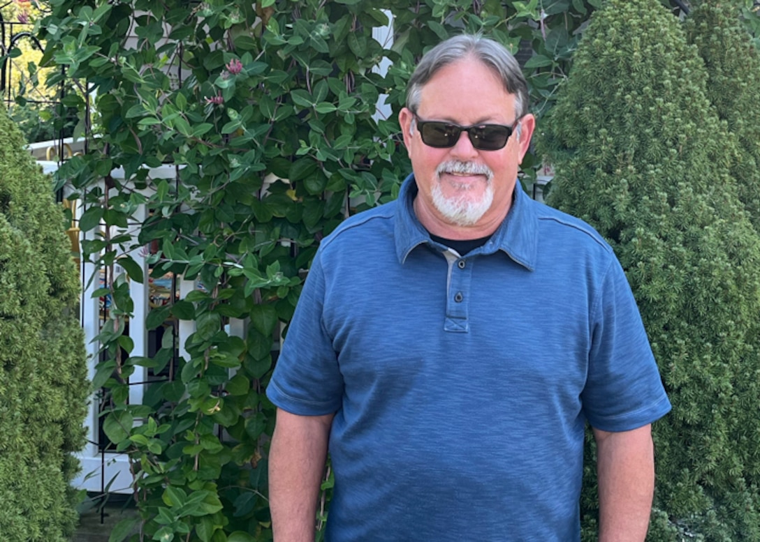 A man in a blue shirt, and dark sunglasses looks at the camera. there is what seems to be a garden or tall green plants behind him.