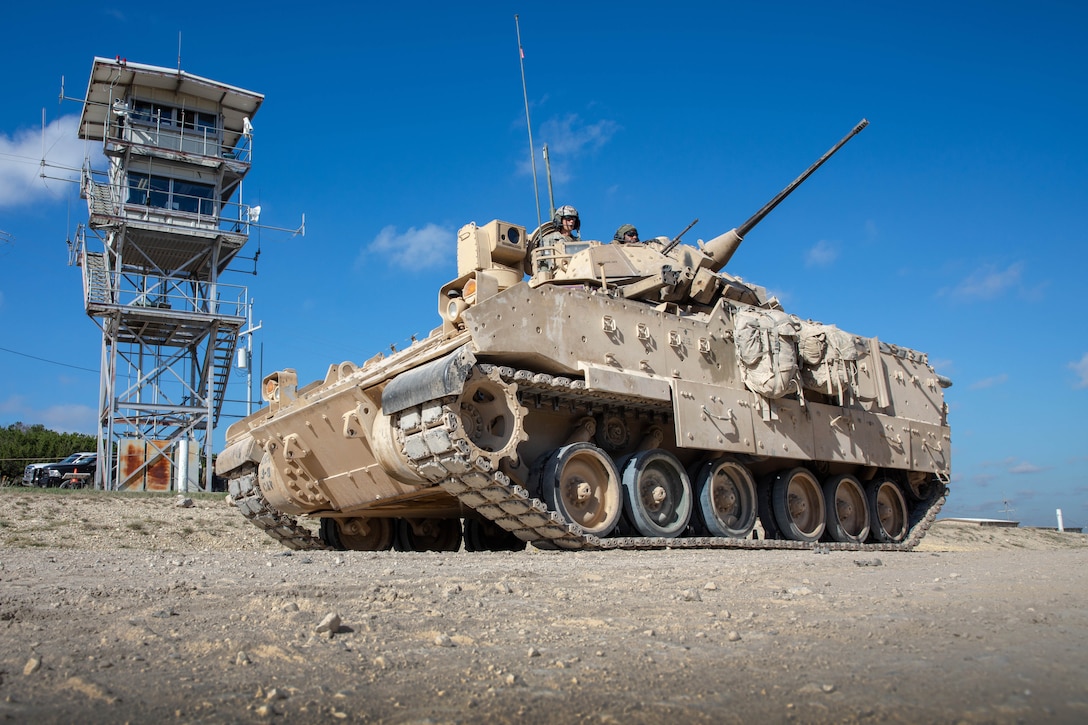 Soldiers operate a tank in front of a satellite tower on a dirt road.