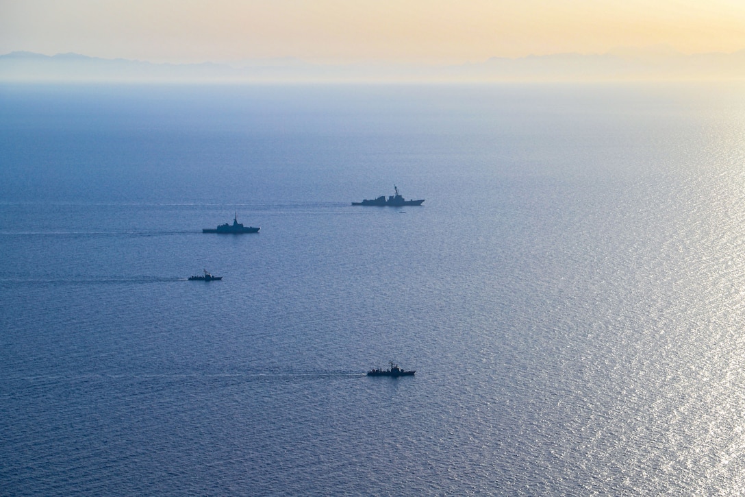 Four ships sail in formation in a body of water as the sun shines on the horizon.