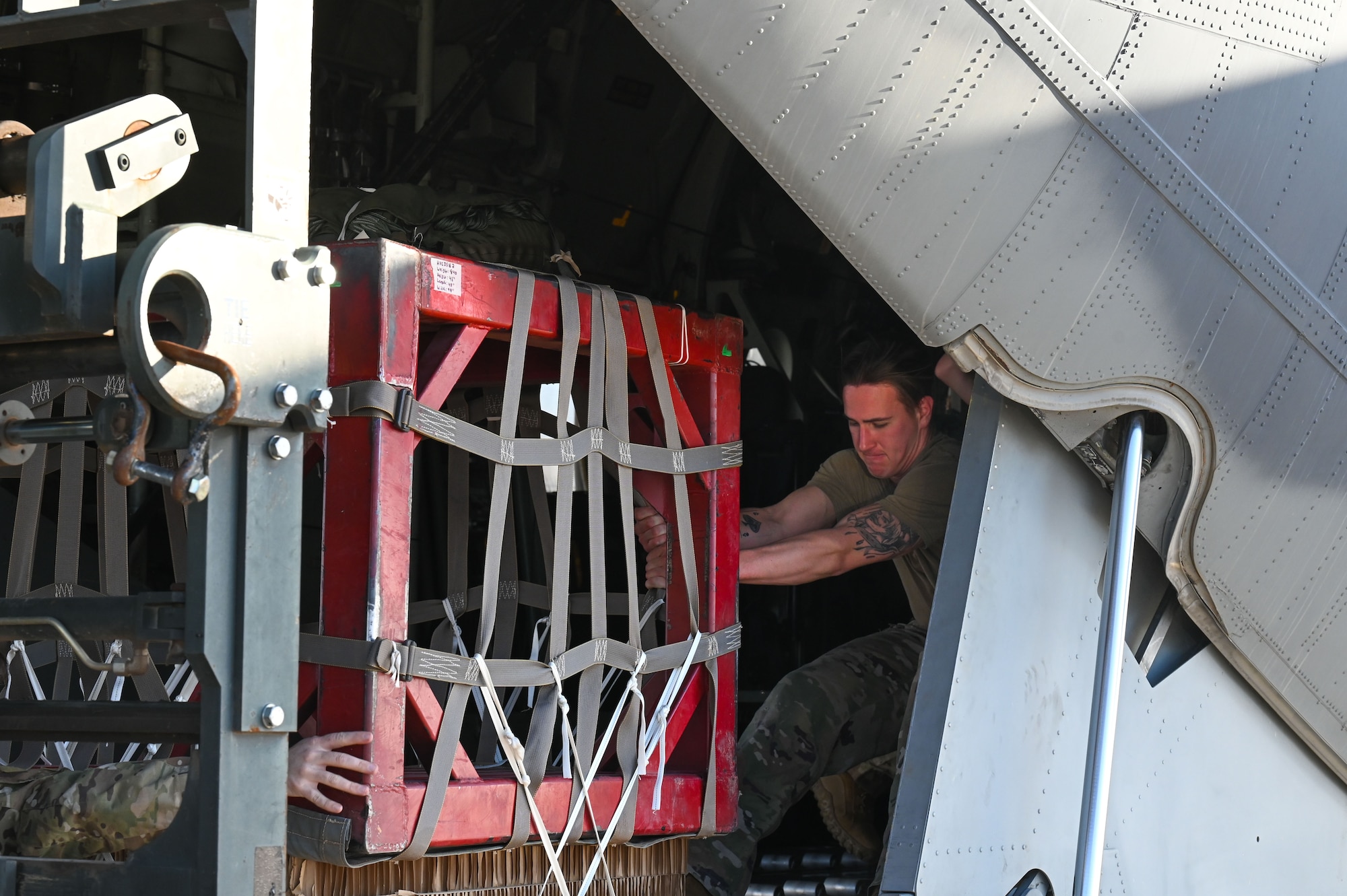 A man in military uniform guides a pallet of cargo onto a military aircraft ramp.