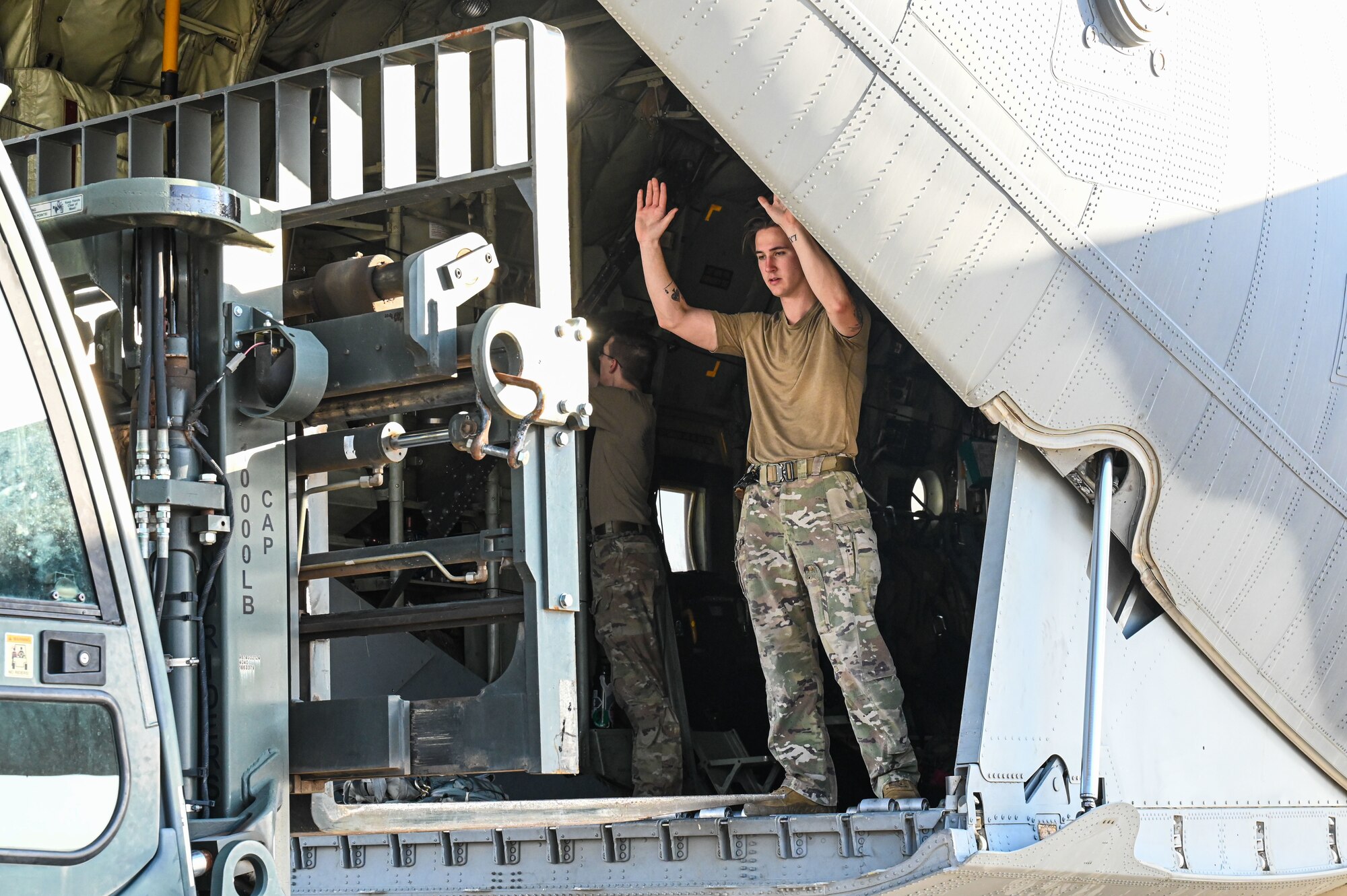 A man in military uniform uses hand signals to guide a fork lift driver delivering cargo onto a military aircraft.
