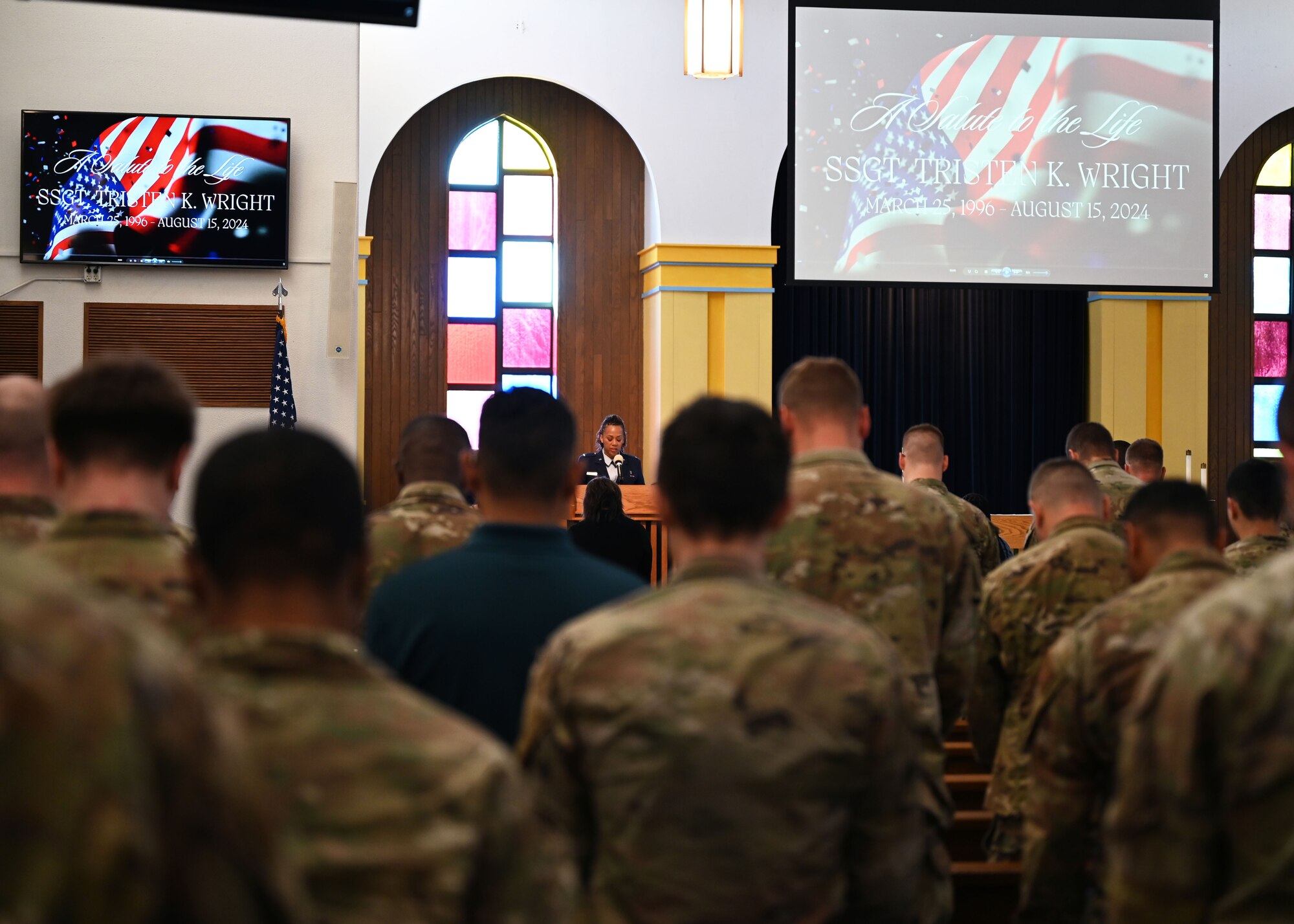A group of men and women in military uniform bow their heads in prayer inside a military chapel. A man in military dress uniform stands at the front of the chapel speaking behind a lectern.