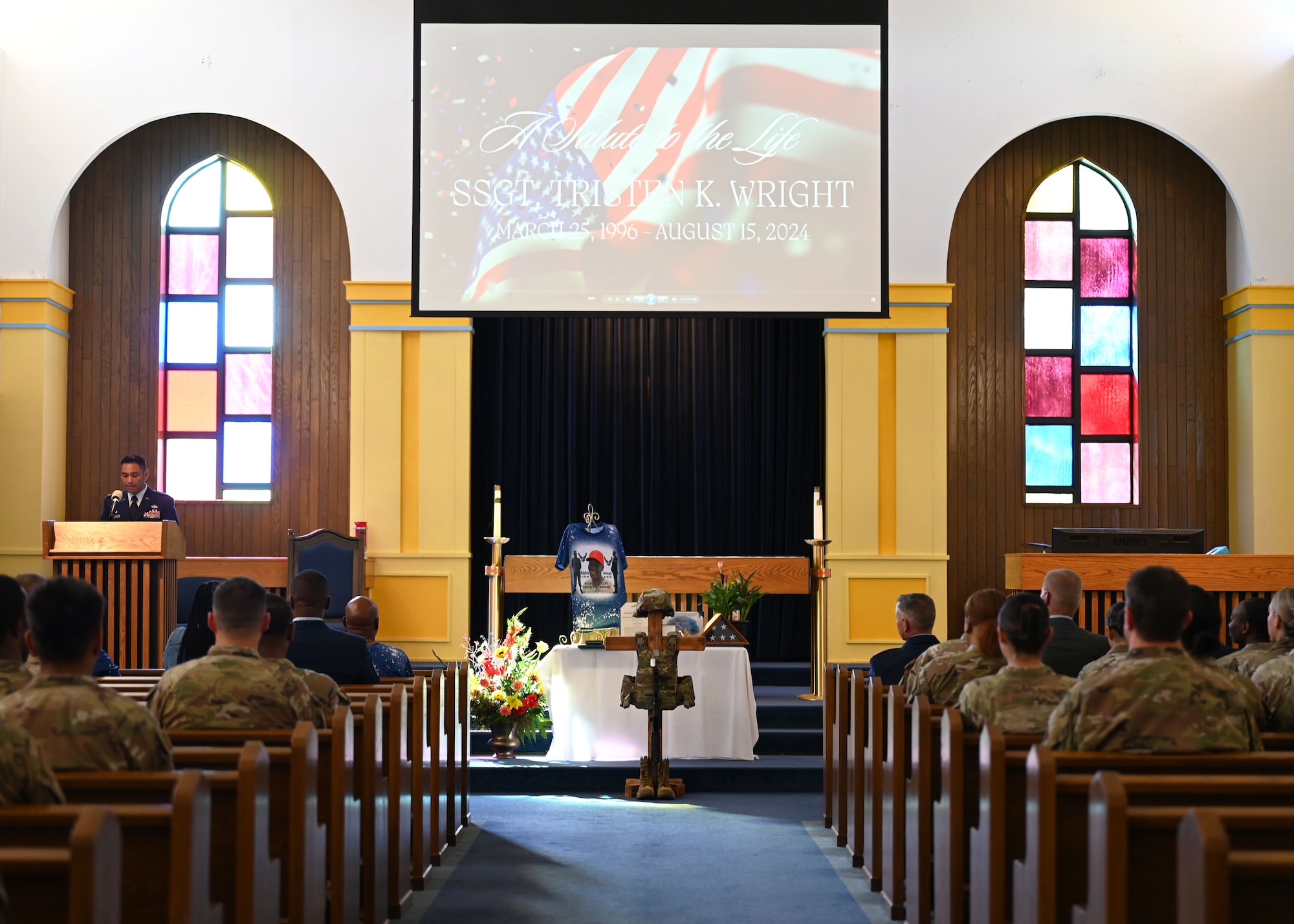 A group of men and women in military uniform sit in the pews of a military chapel during a celebration of life service. The deceased member's personal effects and military equipment are on display at the front of the chapel.