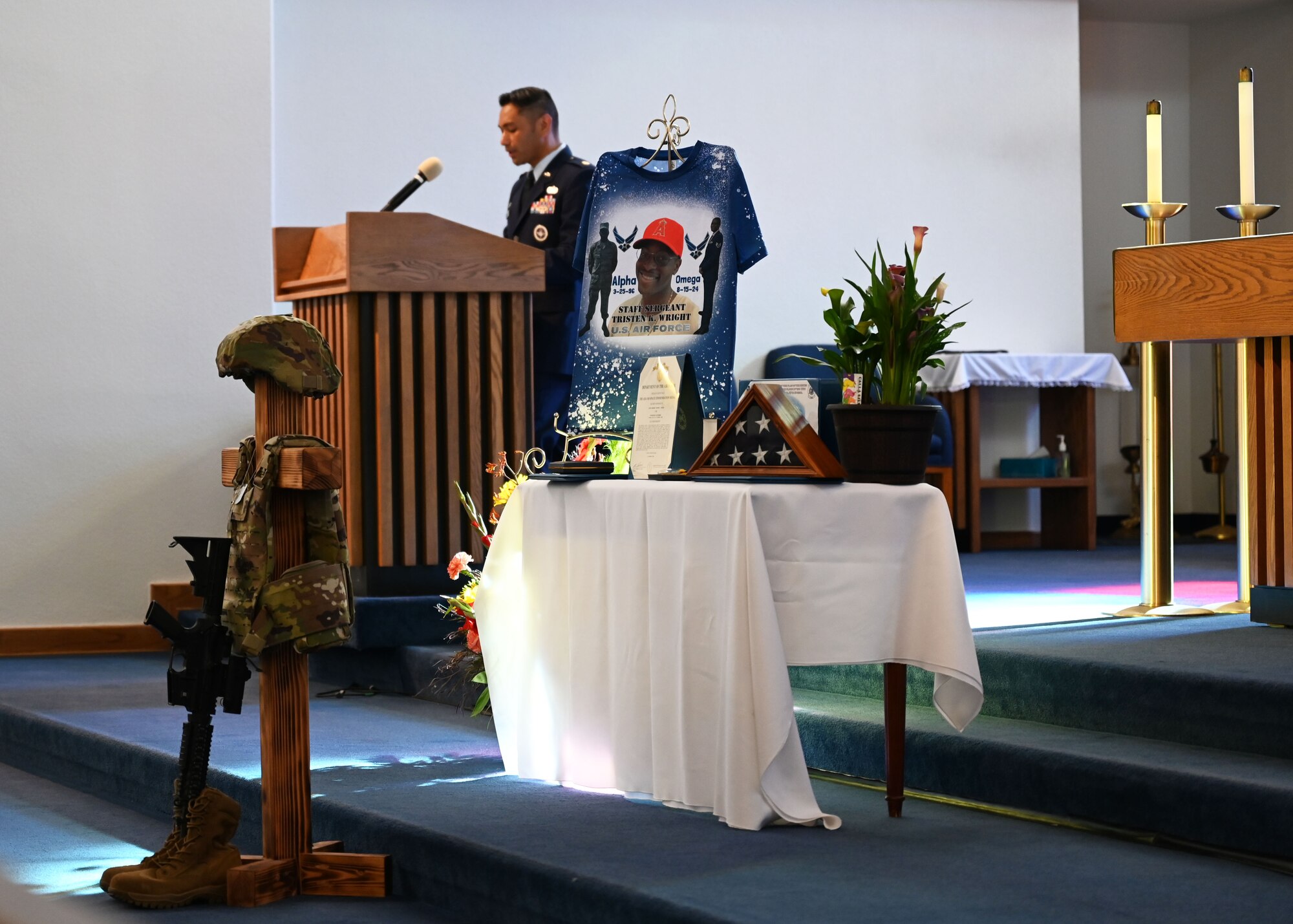 A man in military dress uniform speaks behind a lectern inside a military chapel. A table with military equipment and the personal effects of a deceased member are on display next to the man speaking.