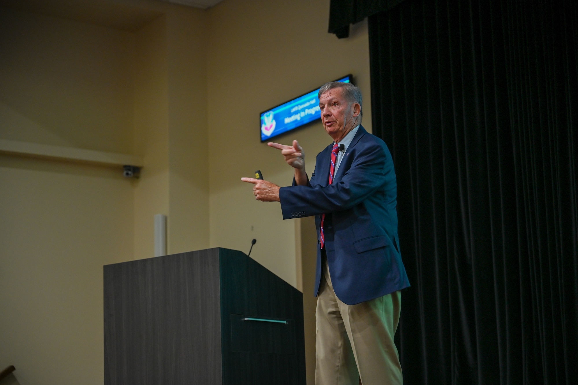 Medium shot of speaker on stage in front of a podium speaking with his fingers pointing in different directions over top of each other.