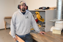 A man poses holding a piece of wood and wearing protective ear covers.