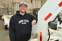 A man in a black pullover hoodie and baseball cap poses against a piece of industrial equipment.