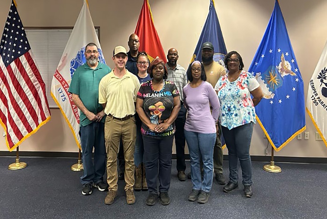 Nine people pose for a group photo in front of the American and military service flags.
