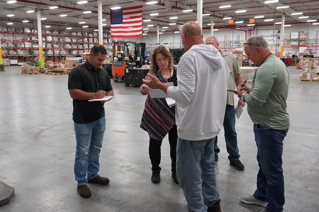 A group of five people stand in a circle inside a distribution warehouse facility taking notes on notepads.