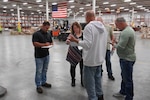 A group of five people stand in a circle inside a distribution warehouse facility taking notes on notepads.