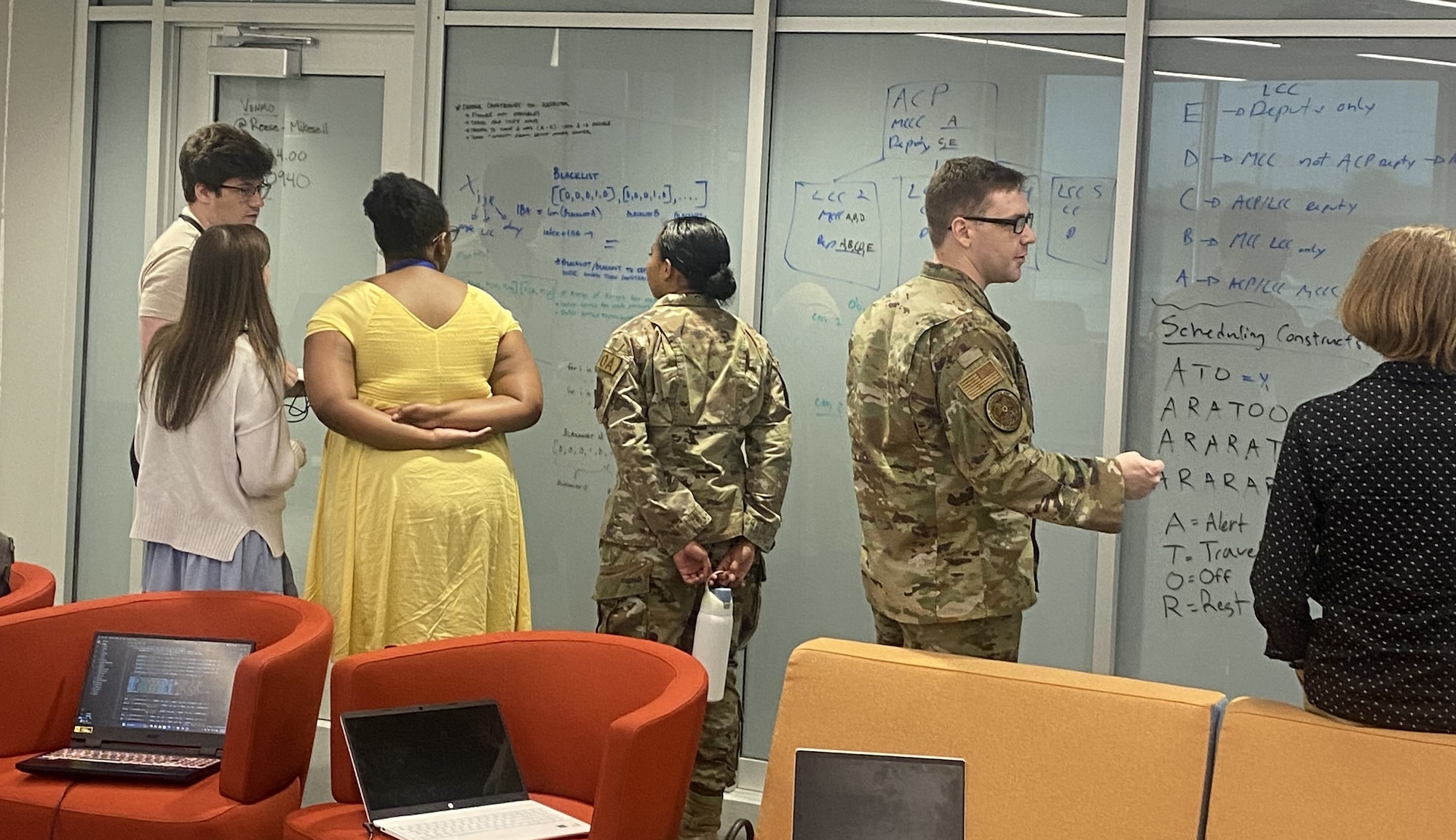 People stand together, facing away from the camera, to look at either each other or the large whiteboards, in an office environment. They are all engaging with one another in their small groups and discussing the contents on the whiteboards.