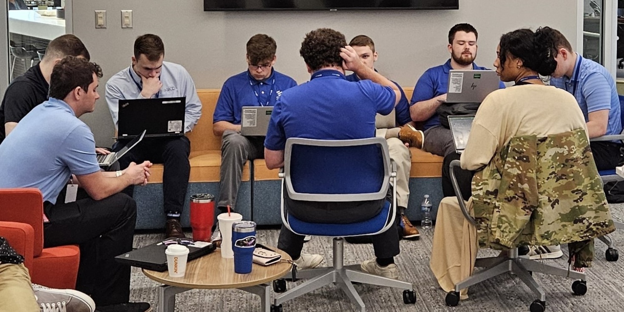 People sit together in an office environment, most have laptops on their laps and all are engaging with one another in their group.