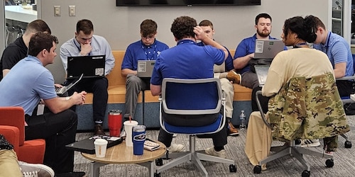 People sit together in an office environment, most have laptops on their laps and all are engaging with one another in their group.