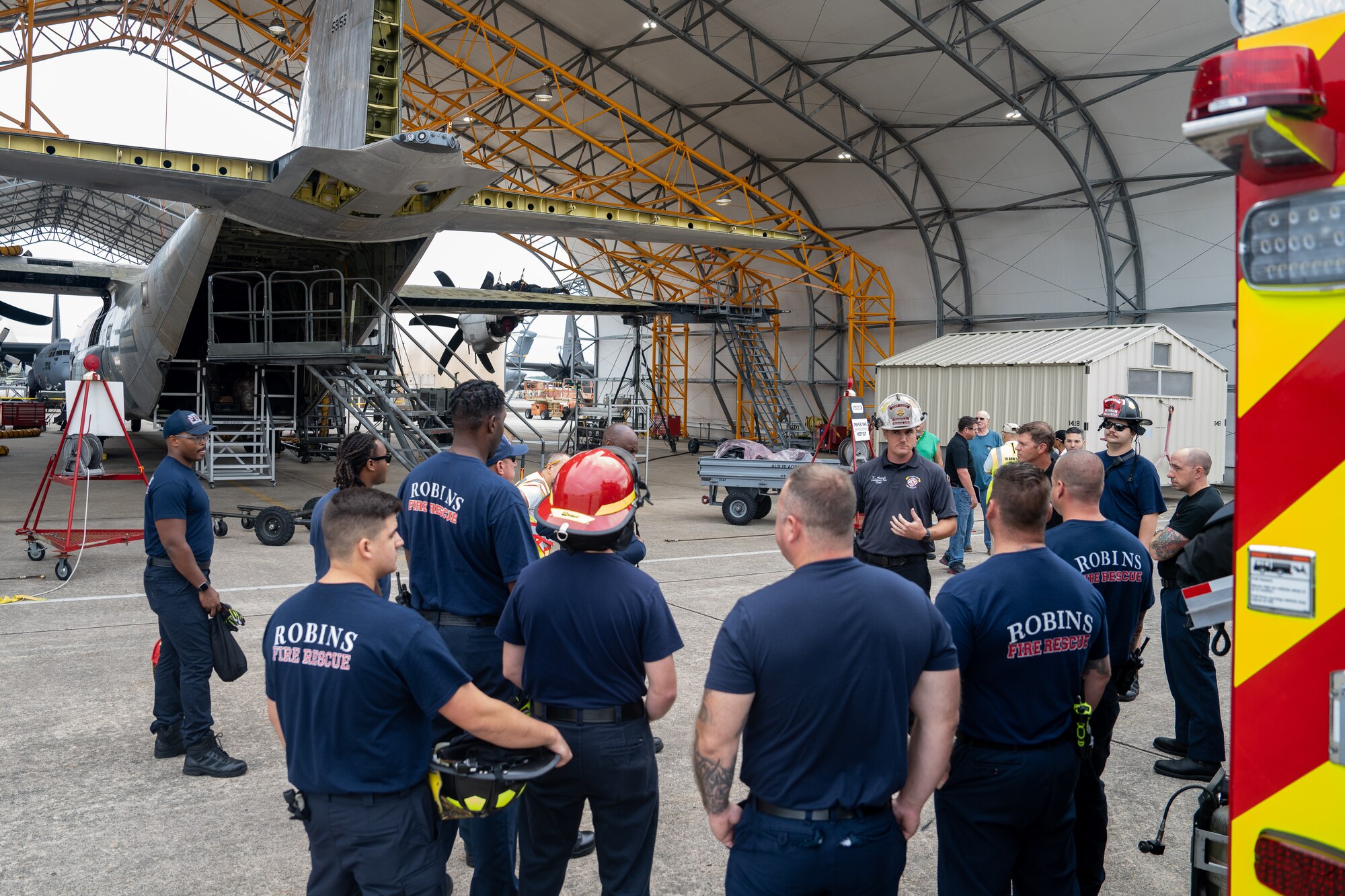 A group of firefighters stand between a fire truck and C-130 Hercules aircraft discussing upcoming training