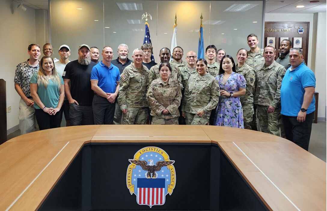 Twenty-three people are posed standing in two rows in a conference room behind a table with the DLA emblem