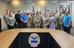 Twenty-three people are posed standing in two rows in a conference room behind a table with the DLA emblem