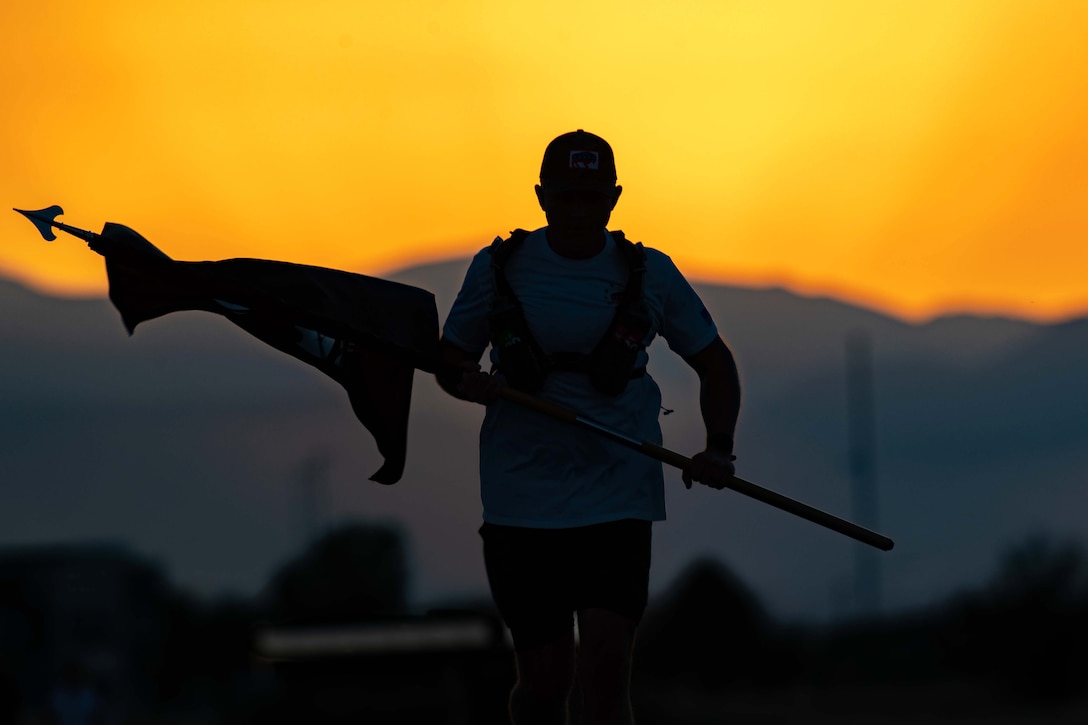 An airman runs while holding a flag in front of yellow orangish sky as seen in silhouette.