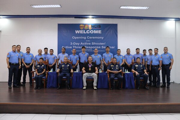 NCIS Special Agent poses, seated, with members of the Manila Police District for posed photo with ceremony banner centered in background.