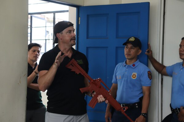 NCIS Special Agent demonstrates use of tactical weapon and methodology while holding prop large capacity weapon as police officers look attentively.