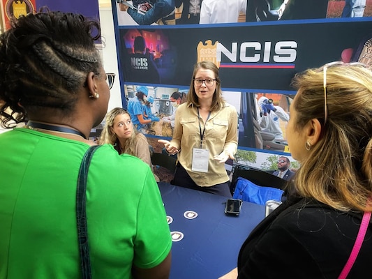 NCIS staff member speaks to attendees at the WIFLE annual conference in front of NCIS recruiting display and table.