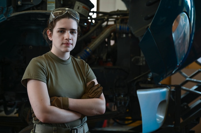 Senior Airman Grace Strobel stands on the left side of the photo with arms crossed staring intently at the camera with a snow removal vehicle in the background