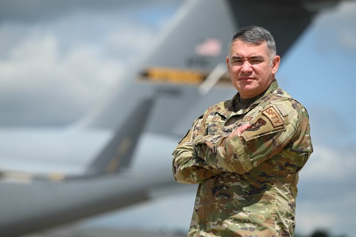 Tech. Sgt. Bradford Scott stands to the right of the photo with arms crossed staring intently at the camera with a C-17 Globemaster III in the background.