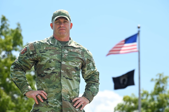 Chief Master Sgt. Dennis Jendrzejewski stands on the left side of the photo with his hands on his hips staring intently at the camera with the American flag waving in the background to the right.