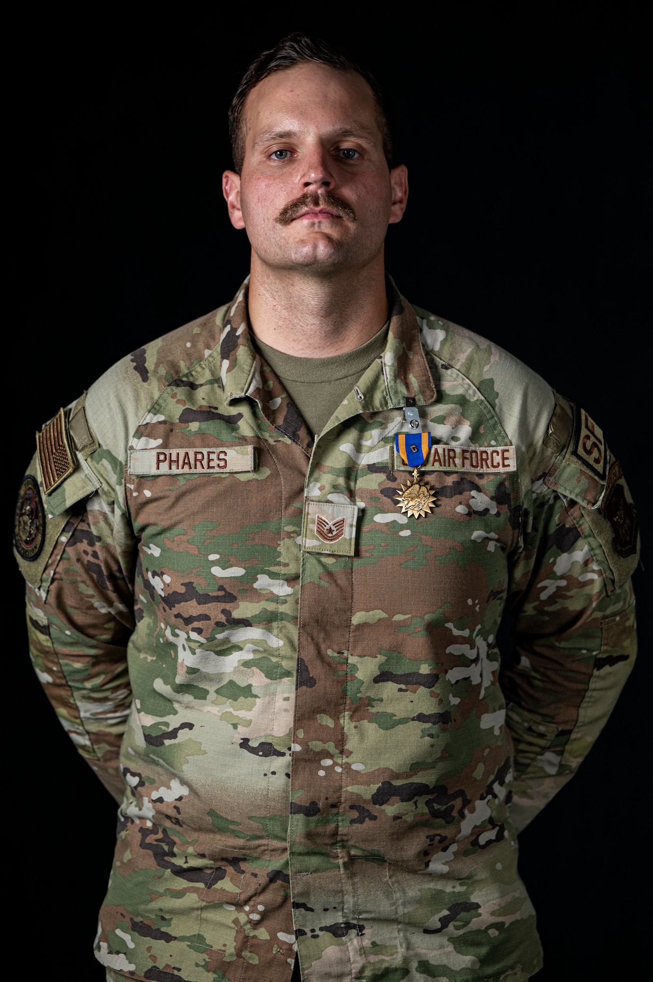 Tech. Sgt. Seth Phares stands mid-frame with his hands behind his back staring intently at the camera with the Air Medal pinned to the left of his chest