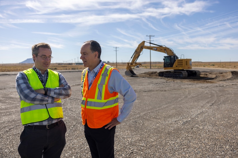 Two people wearing safety vests stand on a dirt field with a backhoe visible in the background.