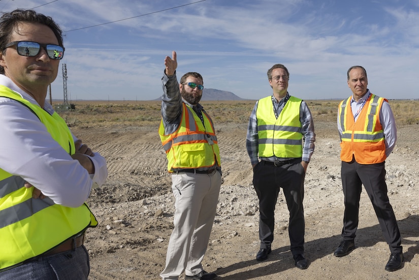 Four people wearing safety vests stand on a dirt field as one person points.