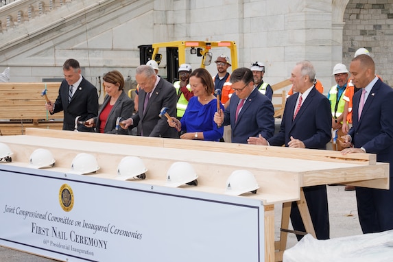 Seven members of Congress are holding hammers and driving nails into a plank of wood. There is sign in front of them that reads First Nail Ceremony.