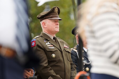 An army general officer in the Army green service uniform is standing with other listening to someone speak during a ceremony.