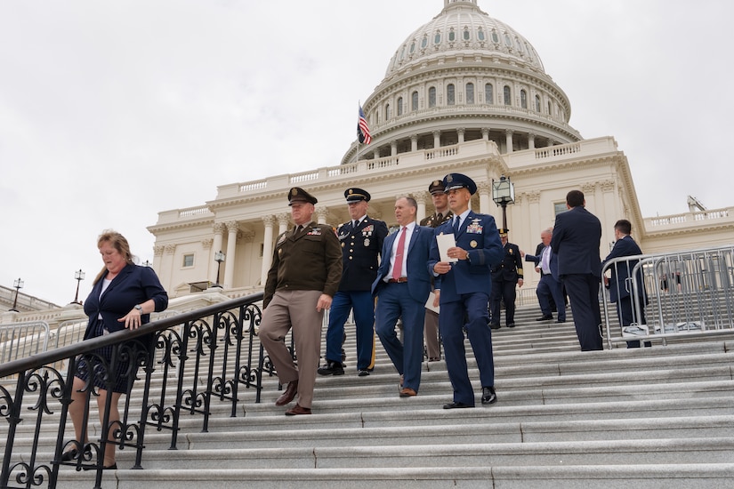 Five men are walking down the steps outside the U.S. Capitol. Four are in military uniforms and one is in a business suit. A woman dressed in a business suit is walking down the steps separately in the foreground.