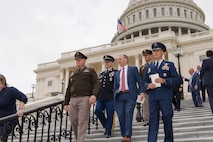 Five men are walking down the steps outside the U.S. Capitol. Four are in military uniforms and one is in a business suit.