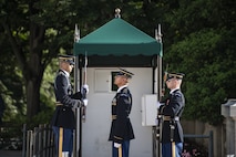 Two soldiers in Army ceremonial uniform (on right) are facing a much taller Army Soldier (on left) who is much taller that the other two Soldiers. They are standing in front of a small, green tent near the Tomb of the Unknown Soldier.