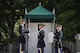 Two soldiers in Army ceremonial uniform (on right) are facing a much taller Army Soldier (on left) who is much taller that the other two Soldiers. They are standing in front of a small, green tent near the Tomb of the Unknown Soldier.