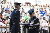 A tall Soldier dressed in dark ceremonial uniform is facing another Soldier who is inspecting his rifle. There is a crowd of people watching on steps nearby.