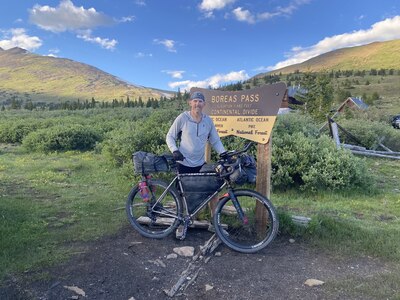 A man stands with his bicycle in front of a large yellow and brown sign that reads "Boreas Pass."  behind him are rolling hills.